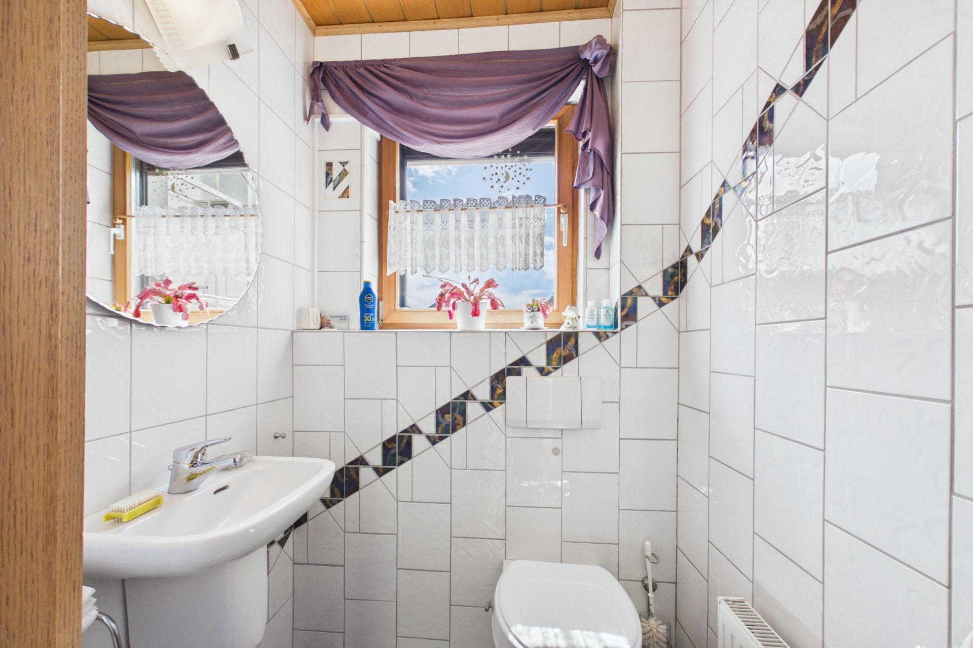 Kleines Bad mit weißen Fliesen, Waschbecken, Toilette und Fenster mit lila Vorhang.