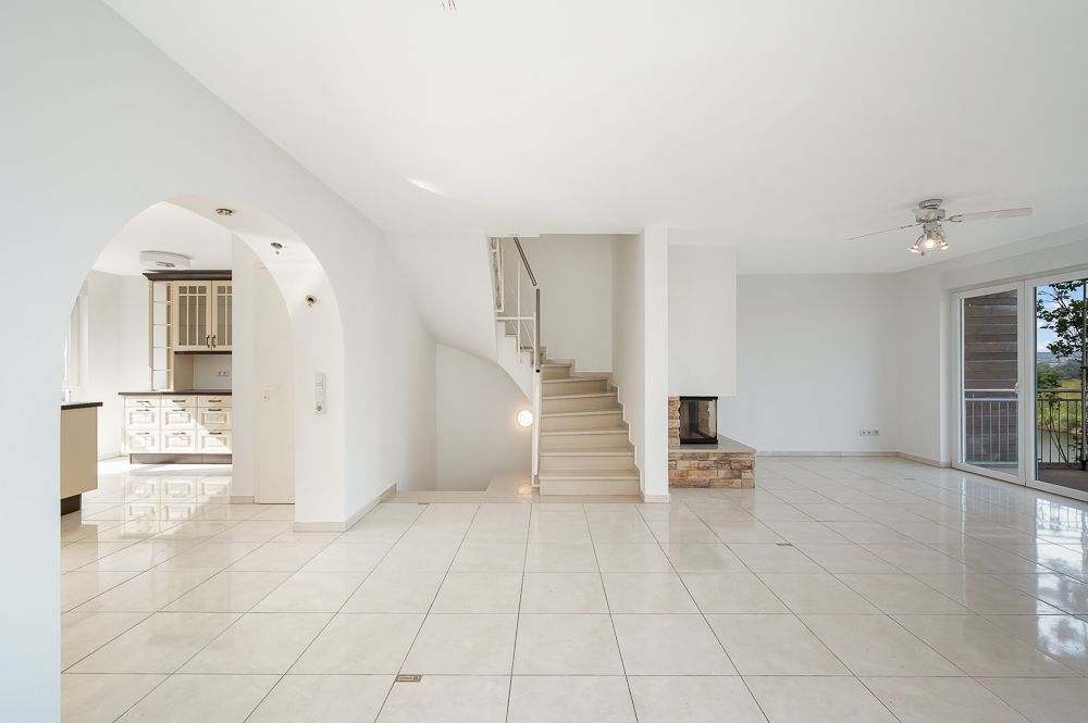 Heller, leerer Wohnraum mit Fliesenboden, Treppe, Kamin und Zugang zur Küche.