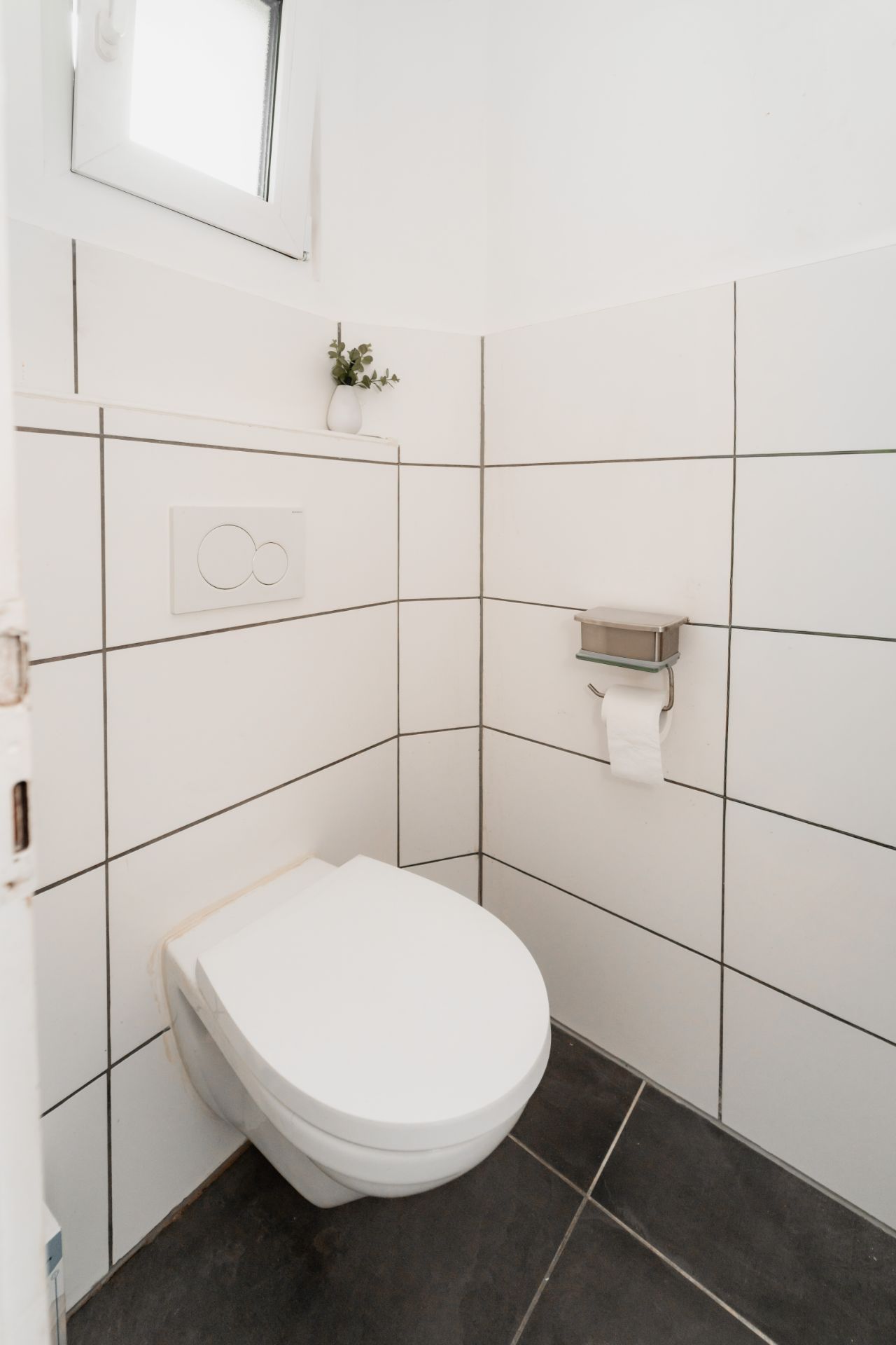 Kleines Badezimmer mit Wandtoilette, Toilettenpapierhalter, weiß gefliest und kleinem Fenster.