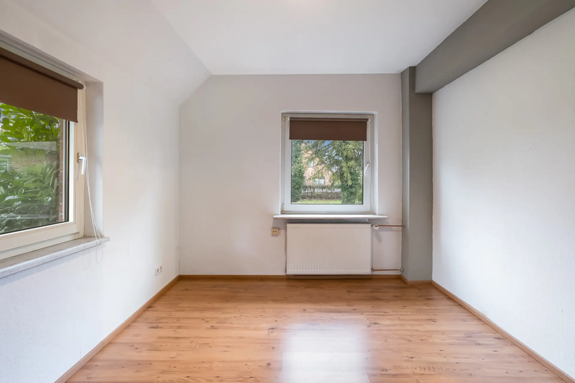 Leerer Raum mit Holzfußboden, zwei Fenstern und einem Heizkörper unter dem mittleren Fenster.