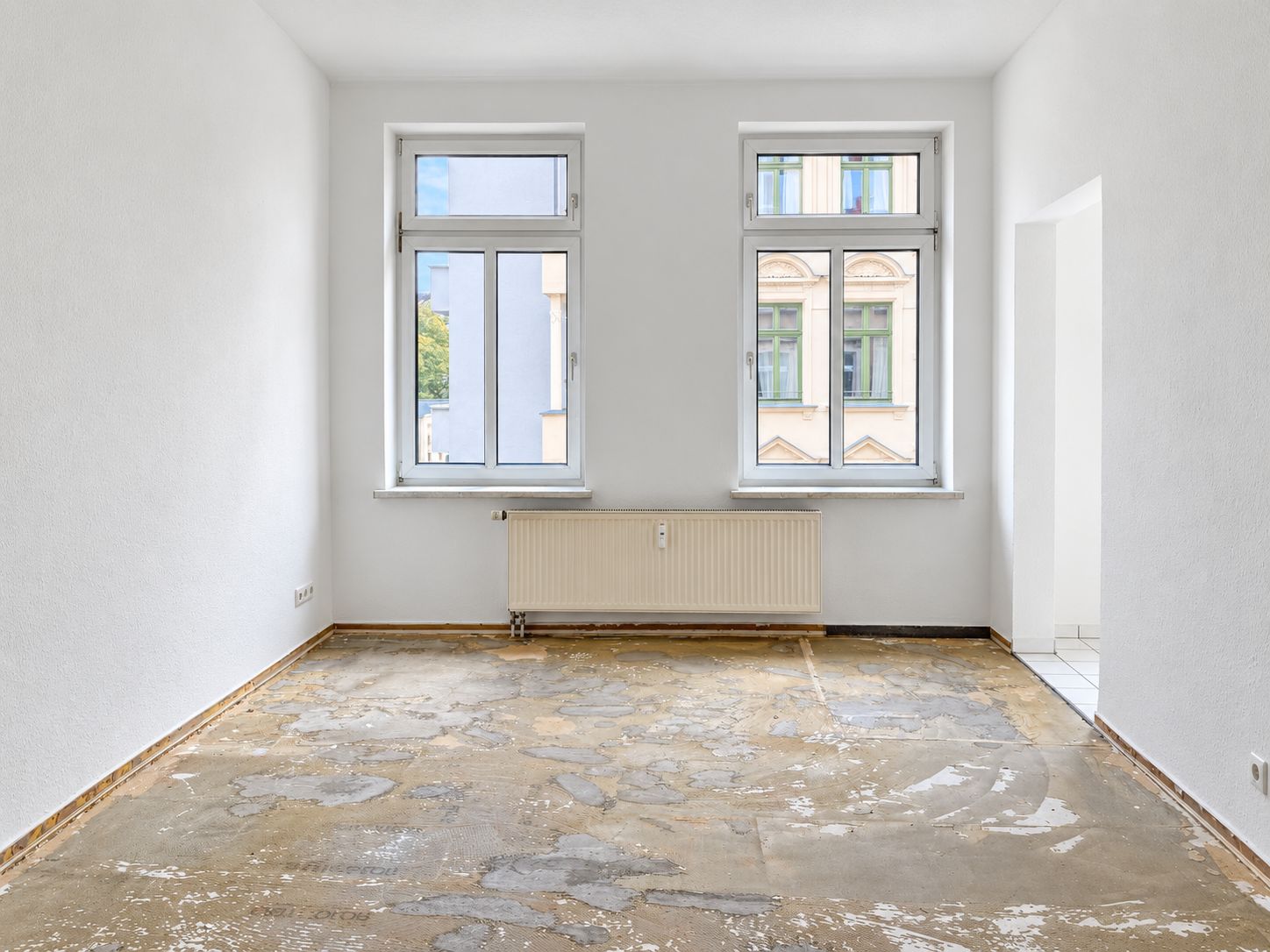Leerer Raum mit zwei Fenstern, Heizung und sichtbar beschädigtem Boden.