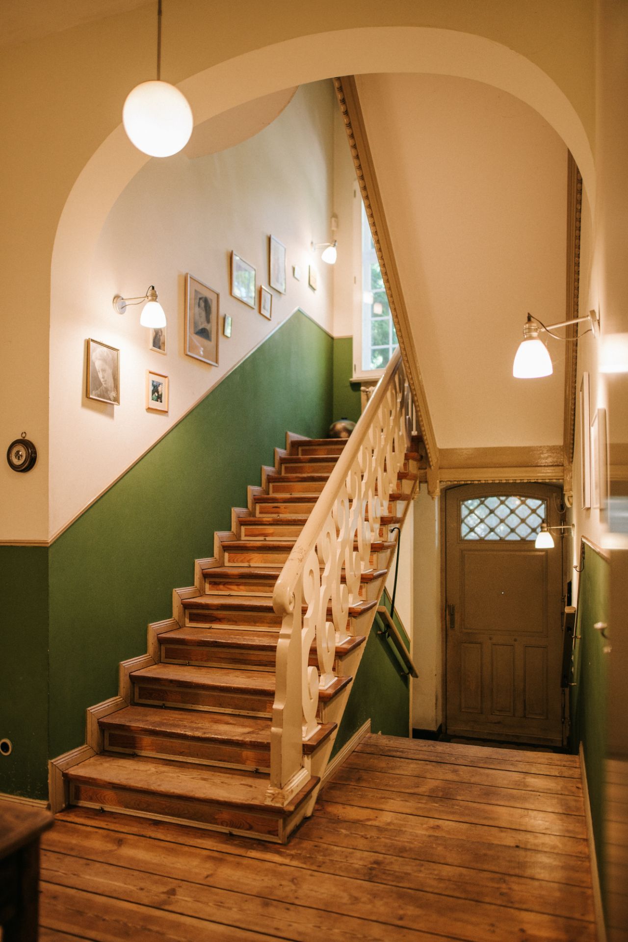 Holztreppe mit grüner Wand, Bildern und Lampen in einem Flur eines Wohnhauses.