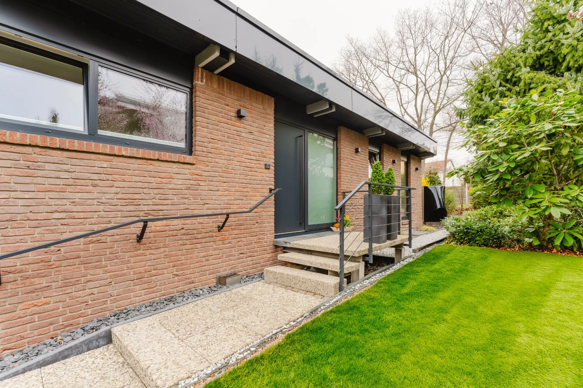 Eingang eines modernen Backsteinhauses mit Treppe, Geländer und gepflegtem Garten.