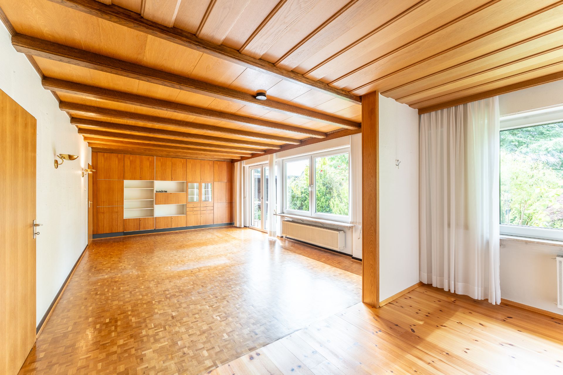 Helles, leeres Wohnzimmer mit Holzboden, Holzdecke und großen Fenstern mit Blick ins Grüne.