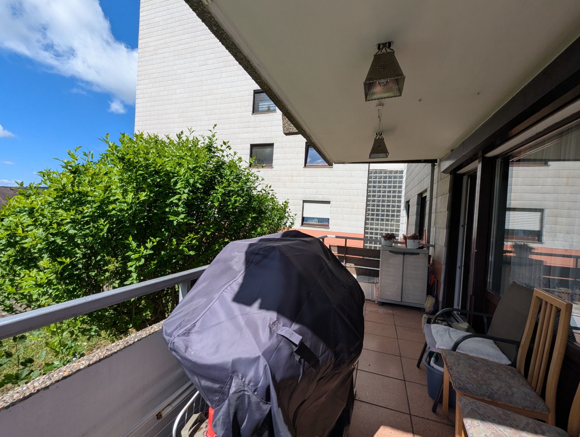 Balkon mit überdachtem Grill, Pflanzen, Stühlen und Blick auf ein Nachbargebäude.