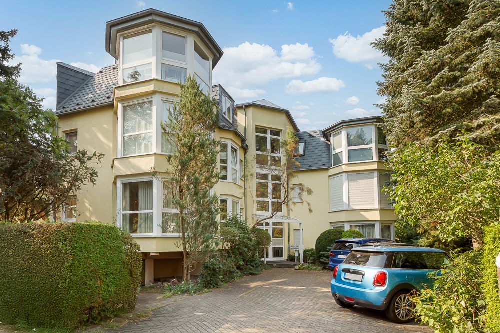 Mehrfamilienhaus mit großen Fenstern, Einfahrt, geparkten Autos und viel Grün im Vordergrund.