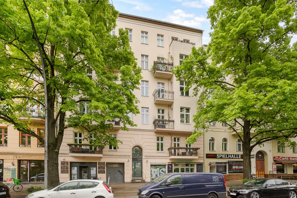 Mehrstöckiges Wohnhaus mit Balkonen, Bäumen und geparkten Autos vor dem Gebäude.
