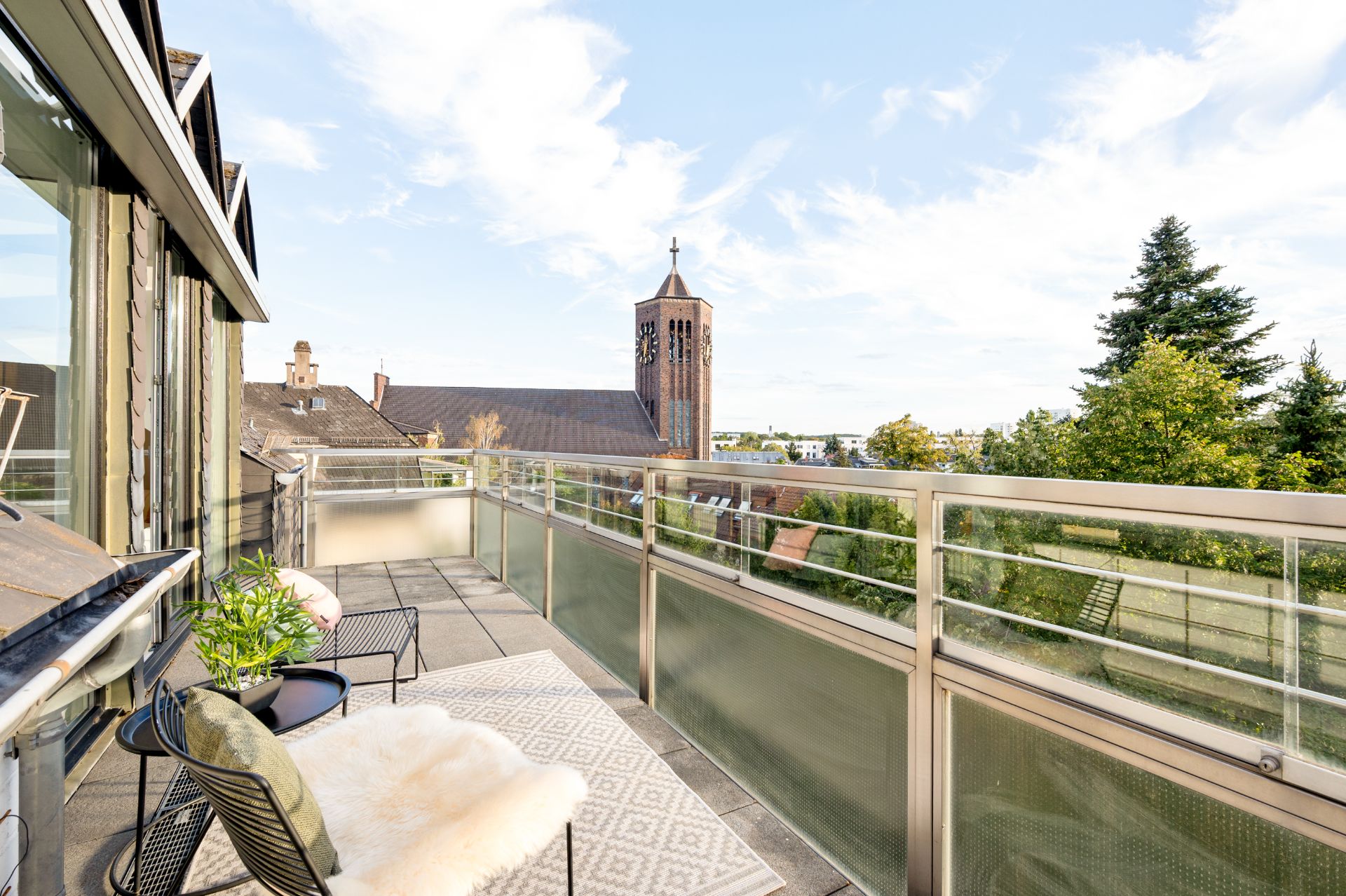Balkon mit Stühlen, Tisch, Teppich und Blick auf Bäume sowie einen Kirchturm im Hintergrund.
