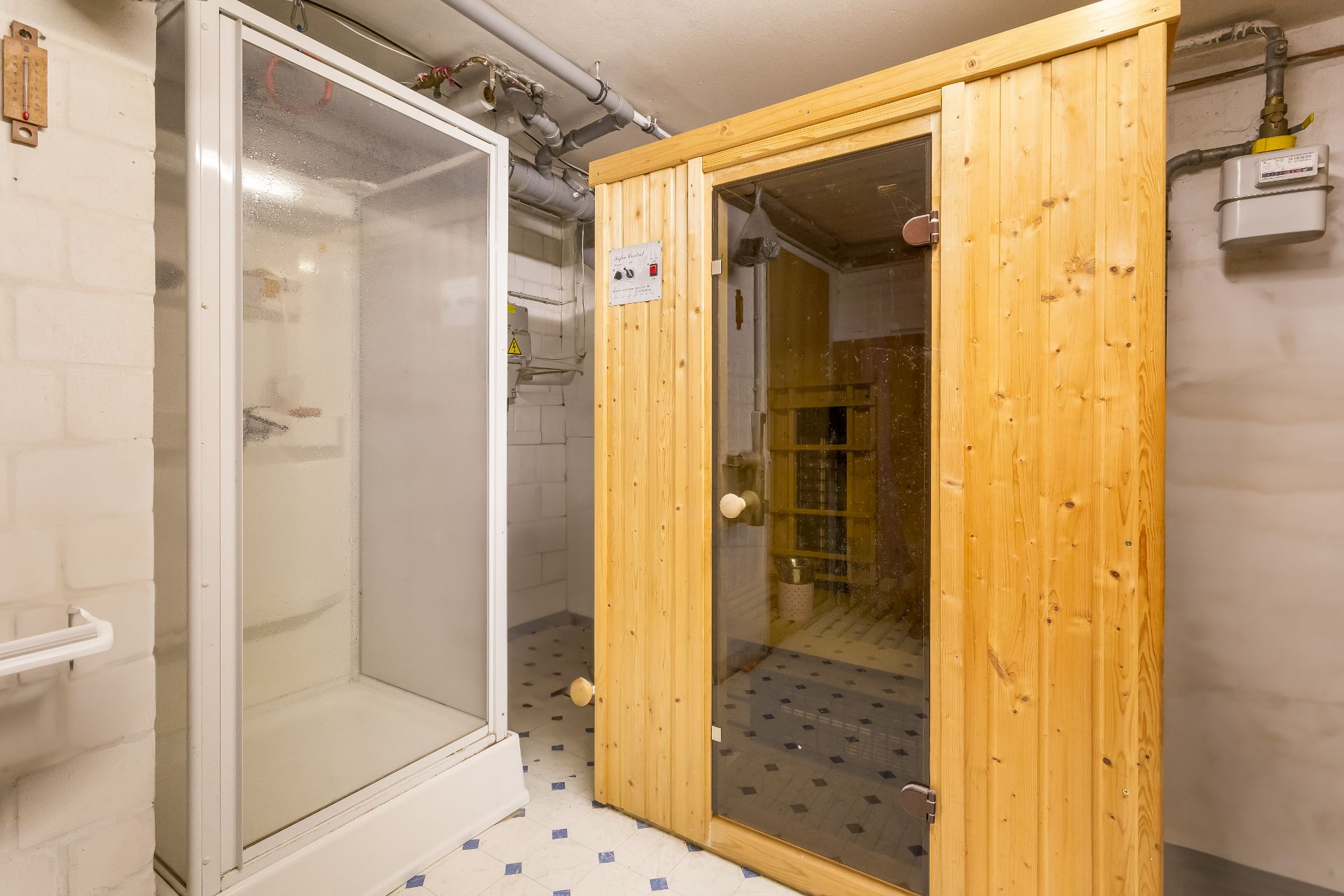 Dusche und Holzsauna in einem Kellerraum mit weißen Wänden und Rohrleitungen.