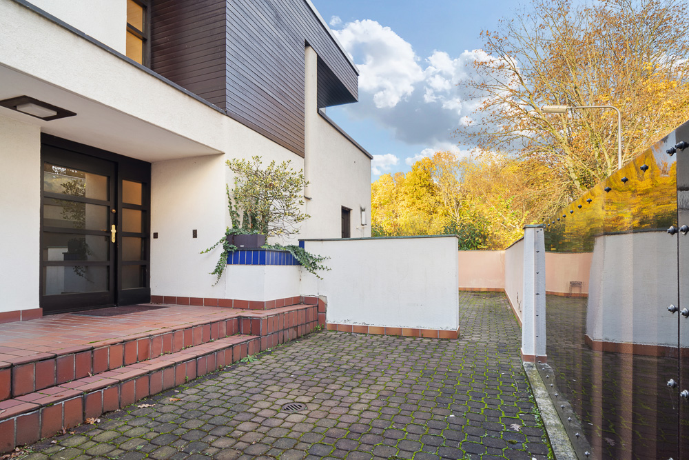 Moderne Hauseingangstreppe mit Pflanzkübel, gepflasterter Weg und Glaszaun, Bäume im Hintergrund.