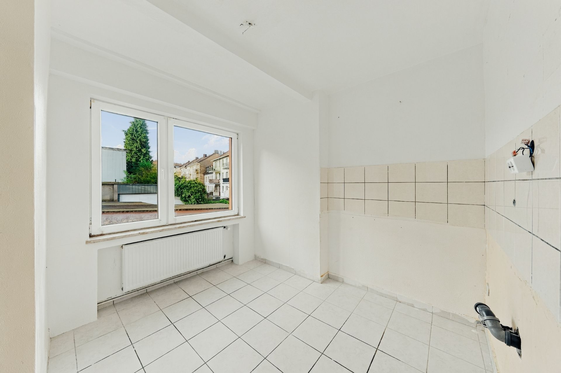 Heller, leerer Raum mit weißen Fliesen, großem Fenster und Heizkörper unter dem Fenster.