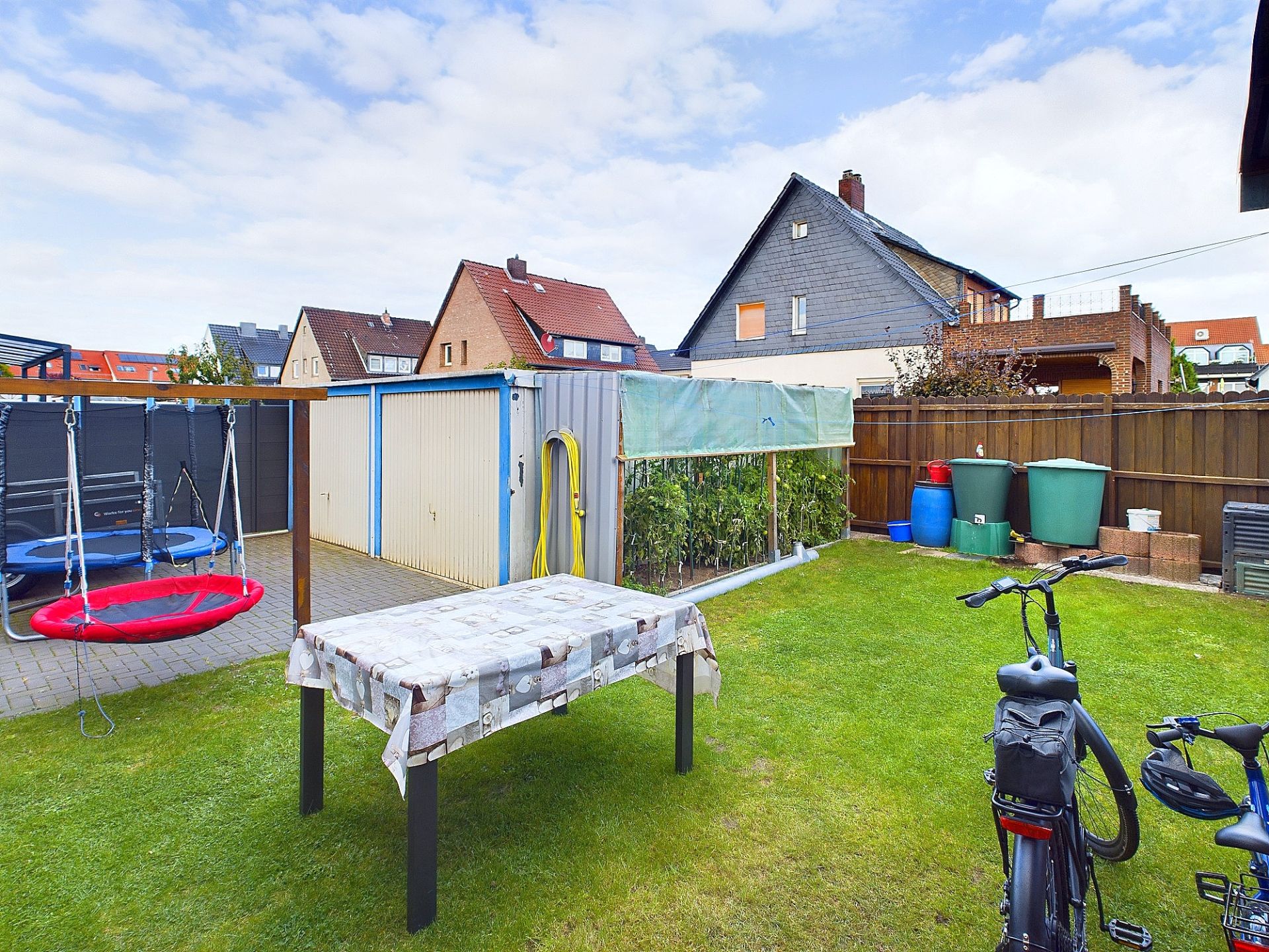 Garten mit Tisch, Schaukeln, Fahrrädern und mehreren Regentonnen neben Schuppen.