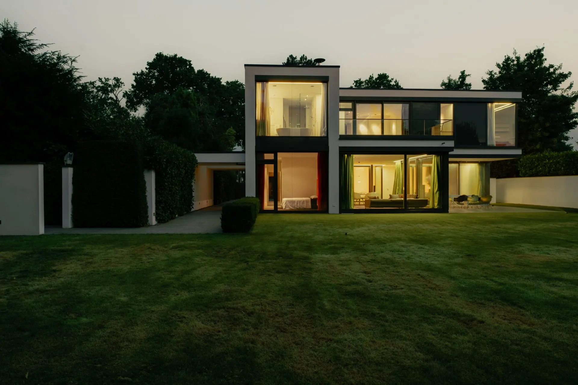 Moderne zweistöckige Villa mit großen Fenstern und beleuchtetem Innenraum, vor einer Rasenfläche.