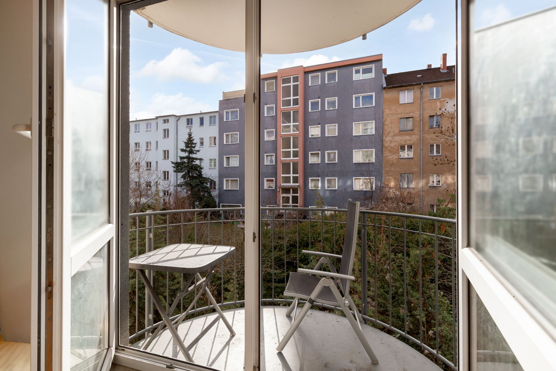 Balkon mit Tisch und zwei Stühlen, Blick auf Wohngebäude und grüne Bäume.