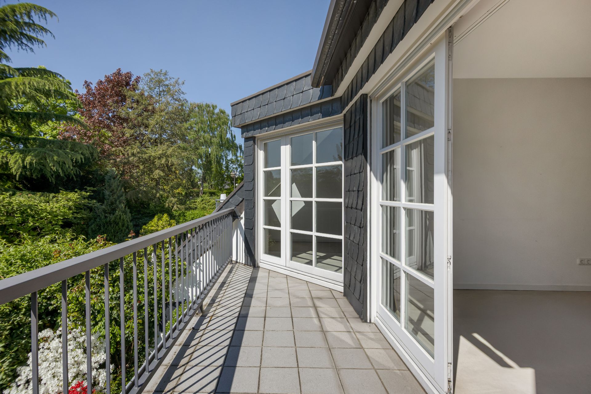 Heller Balkon mit Fliesenboden und Geländer, daneben große Fenster, Blick ins Grüne.