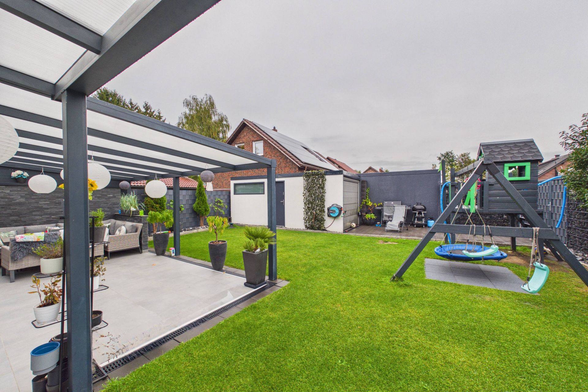 Garten mit überdachter Terrasse, Rasenfläche, Spielgerüst mit Schaukel und Rutsche.