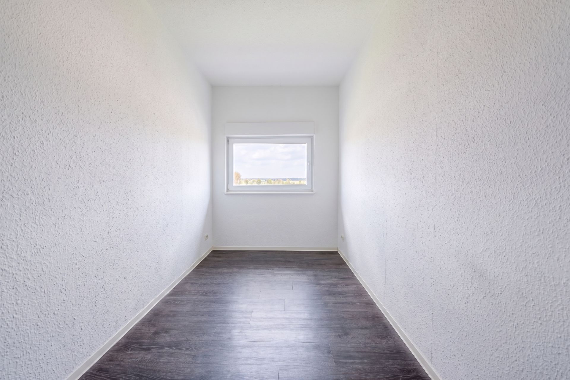 Schmaler, leerer Raum mit weiß gestrichenen Wänden, dunklem Boden und einem Fenster am Ende.