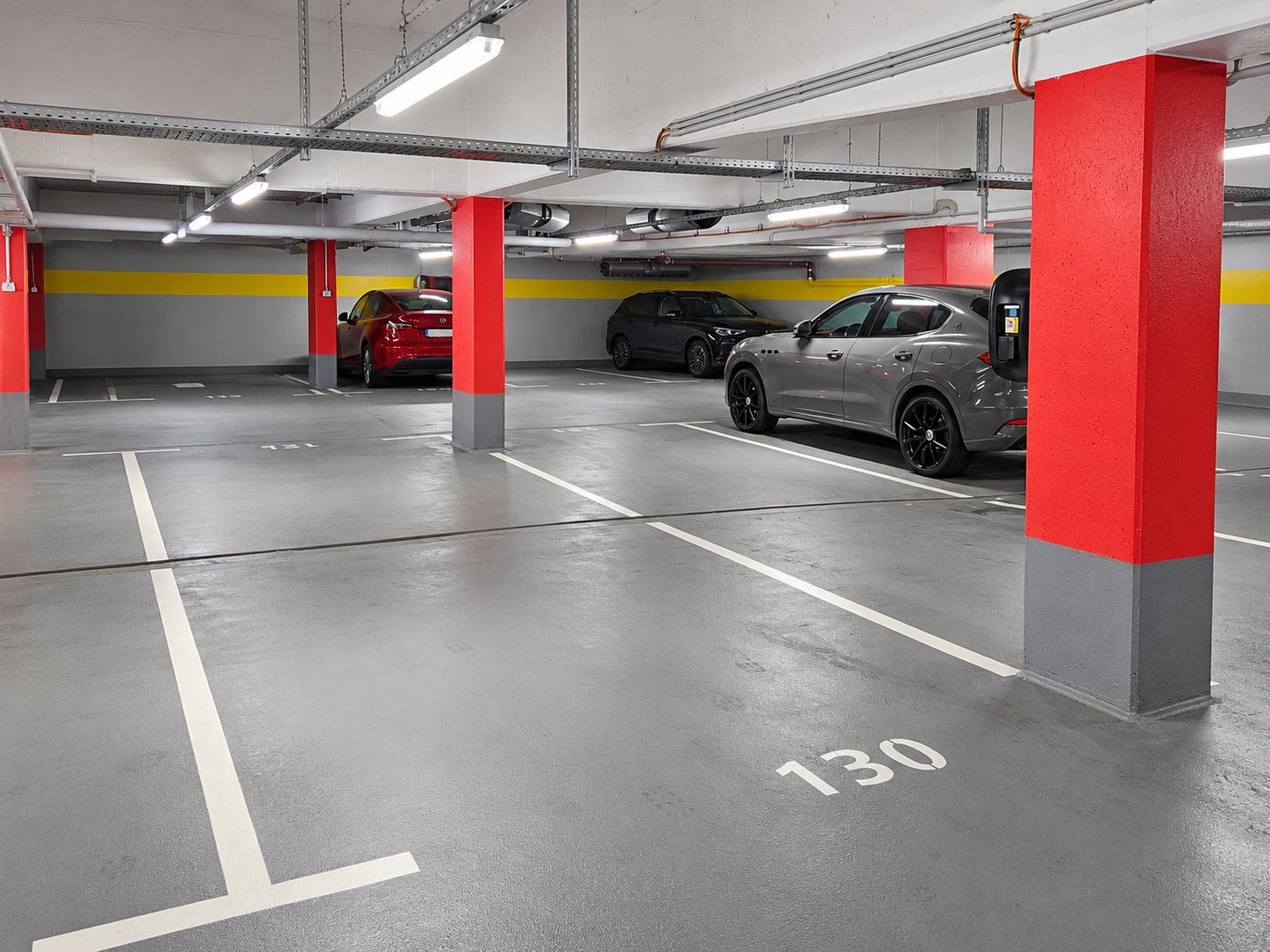 Parkhaus mit markierten Stellplätzen, rot-grauen Säulen und einigen geparkten Autos.