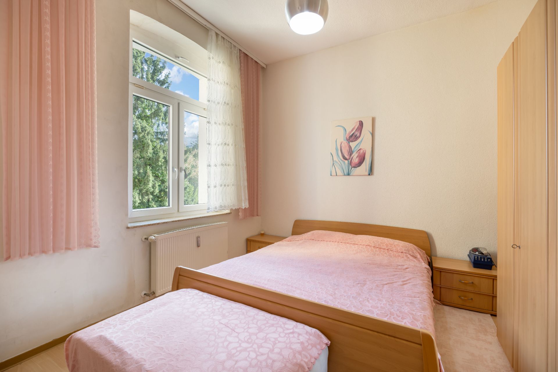 Helles Schlafzimmer mit Doppelbett, rosa Bettwäsche, großem Fenster und Kleiderschrank.