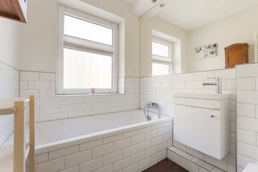 Helles Badezimmer mit Badewanne, Waschbecken, großem Spiegel und zwei Fenstern.