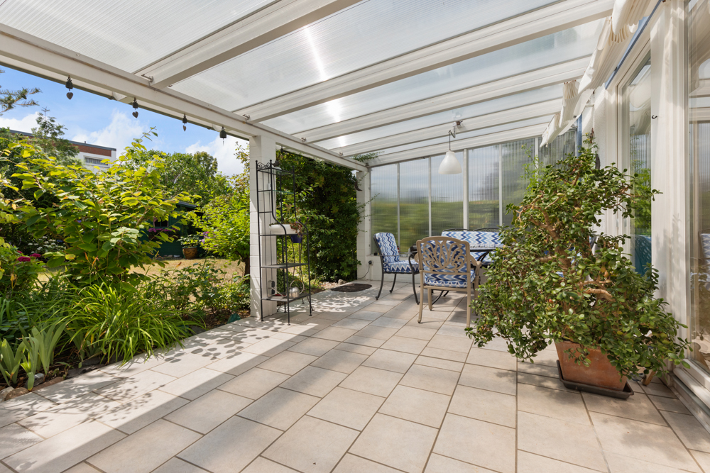 Überdachte Terrasse mit Gartenmöbeln, großen Pflanzen und Blick ins Grüne.