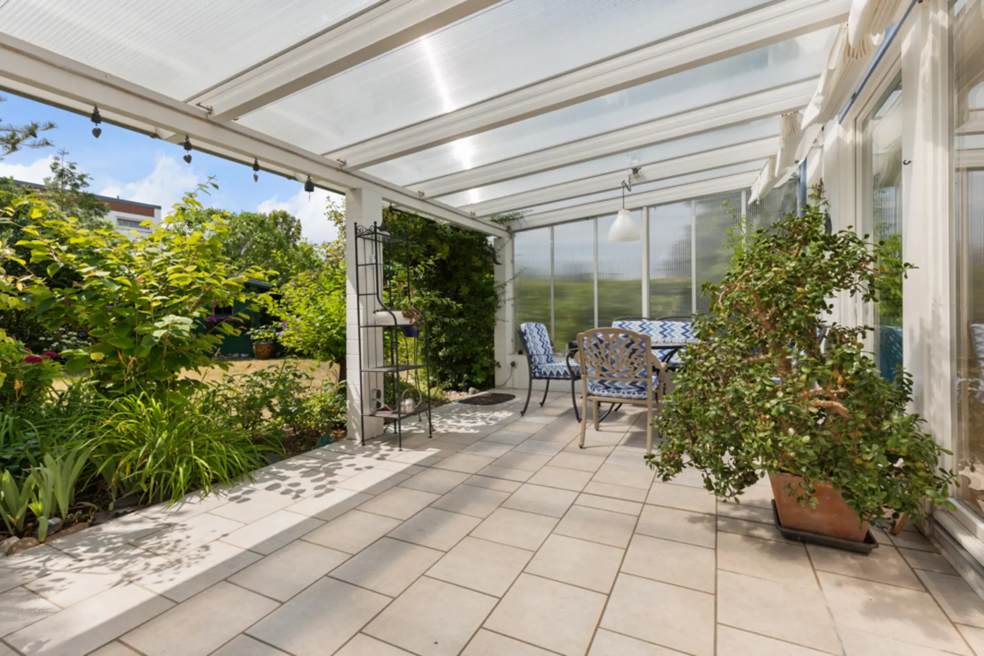 Überdachte Terrasse mit Gartenmöbeln, großen Pflanzen und Blick ins Grüne.