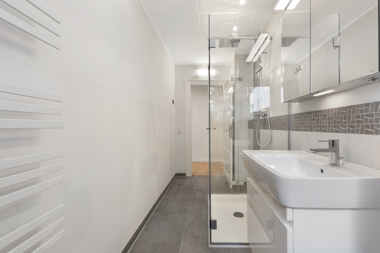 Modernes Bad mit Dusche, großem Waschbecken und Spiegeln, graue Fliesen und Handtuchheizkörper.