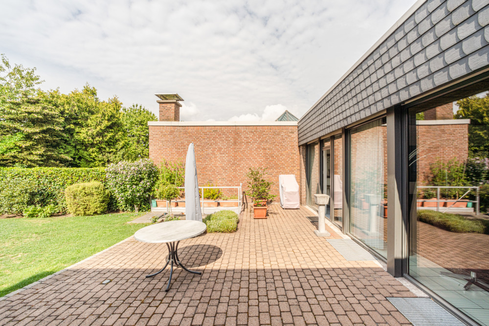 Sonnige Terrasse mit Tisch, Sonnenschirm und Pflanzen, neben einem modernen Haus mit großen Fenstern.