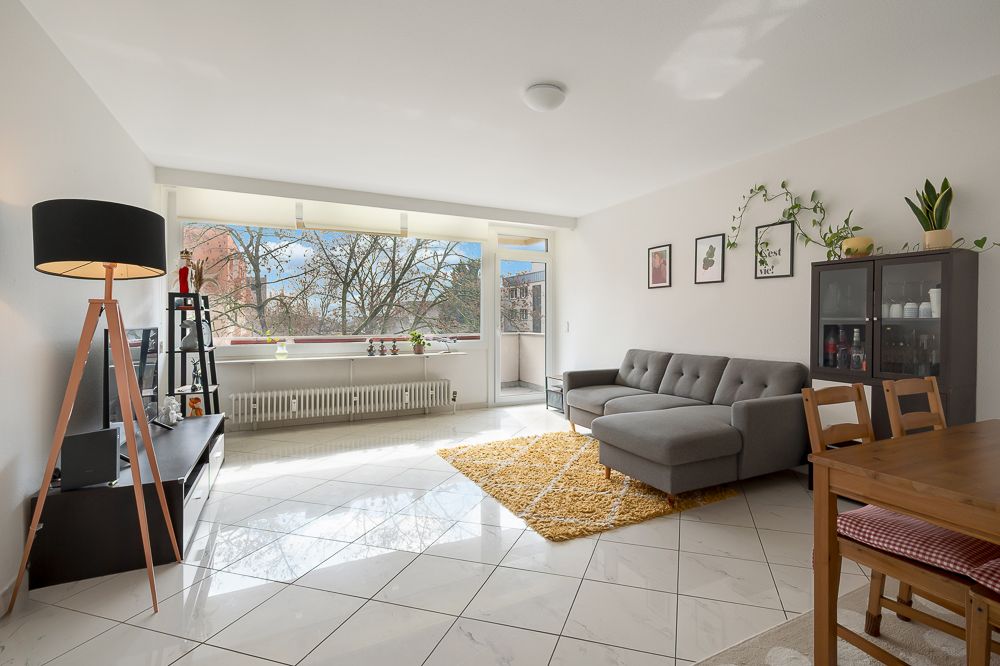 Helles Wohnzimmer mit Sofa, Esstisch, Regal, Stehlampe und großem Fenster mit Blick ins Grüne.