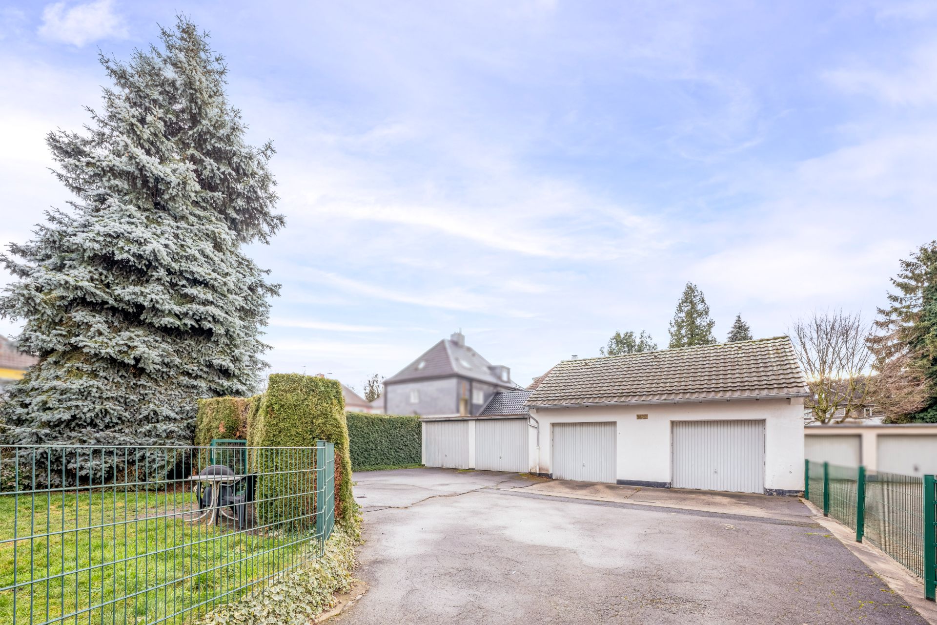 Drei Garagen mit weißen Toren neben einem Garten mit Zaun und großer Tanne.
