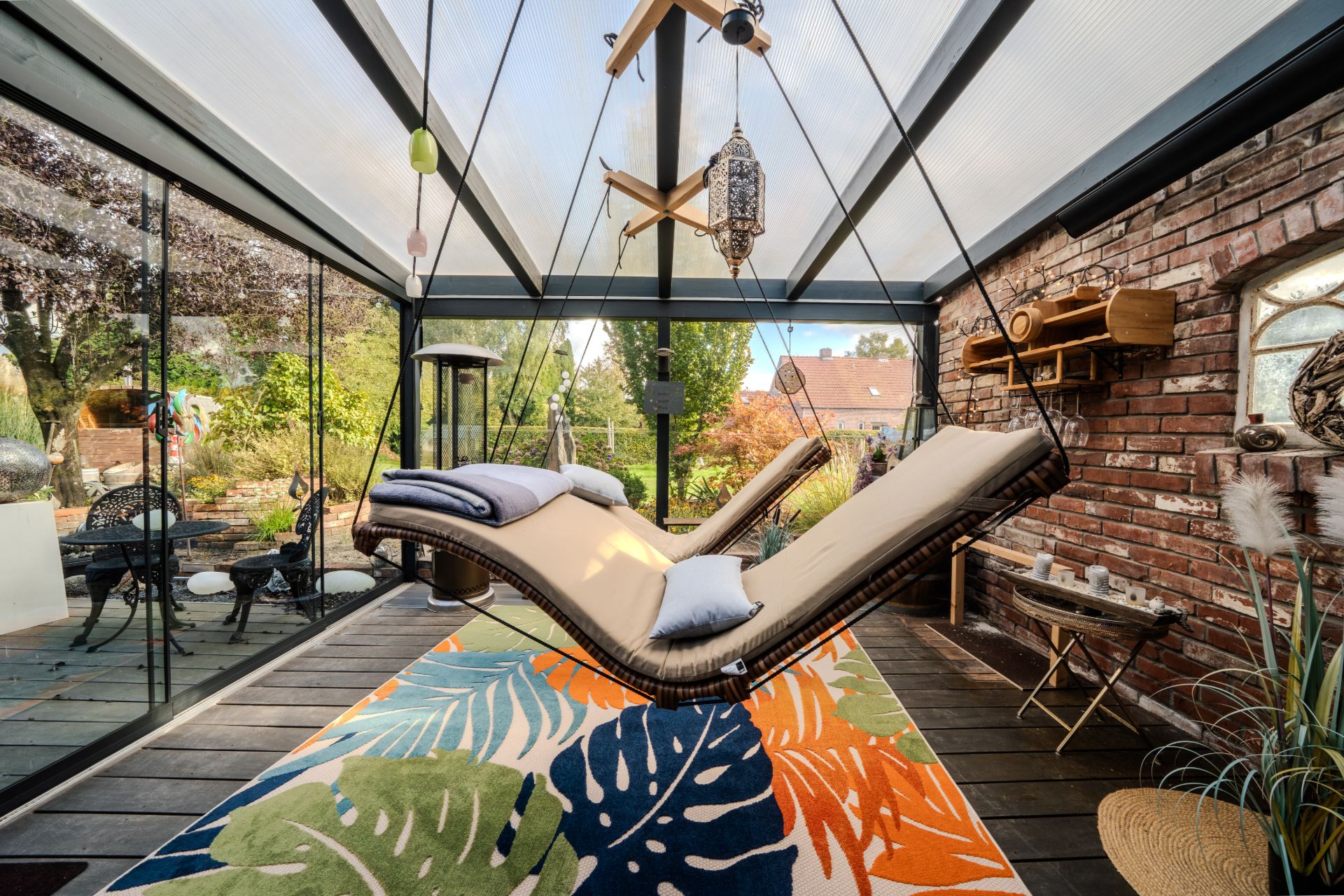 Hängeliege in verglastem Wintergarten mit buntem Teppich und Brickwand.