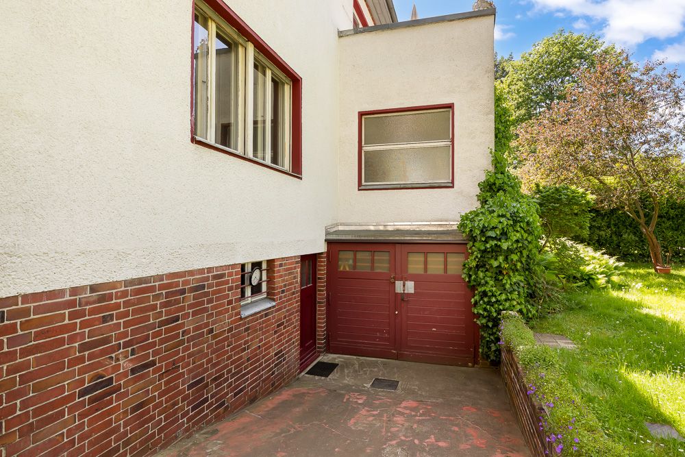 Garage mit braunen Toren an einem Wohnhaus, daneben grüner Garten mit Bäumen.