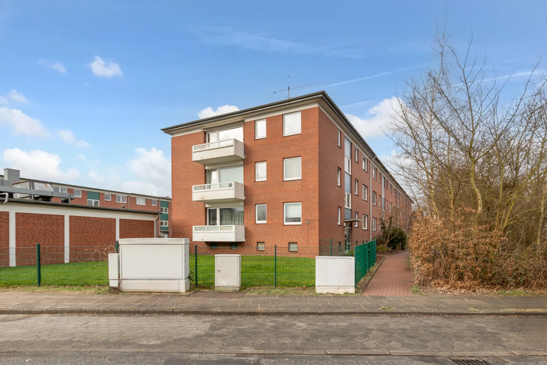 Mehrstöckiges Wohnhaus aus rotem Backstein mit Balkonen und grünem Vorgarten.