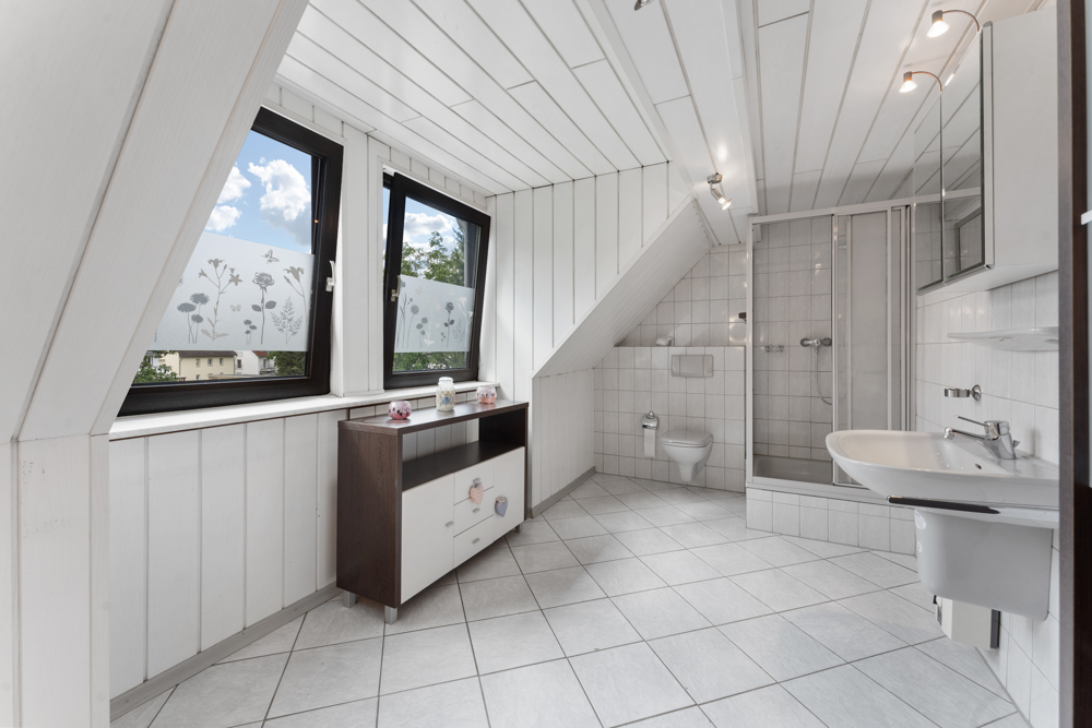 Helles Badezimmer mit Dusche, WC, Waschbecken und zwei Fenstern mit Blumenmuster-Folie.