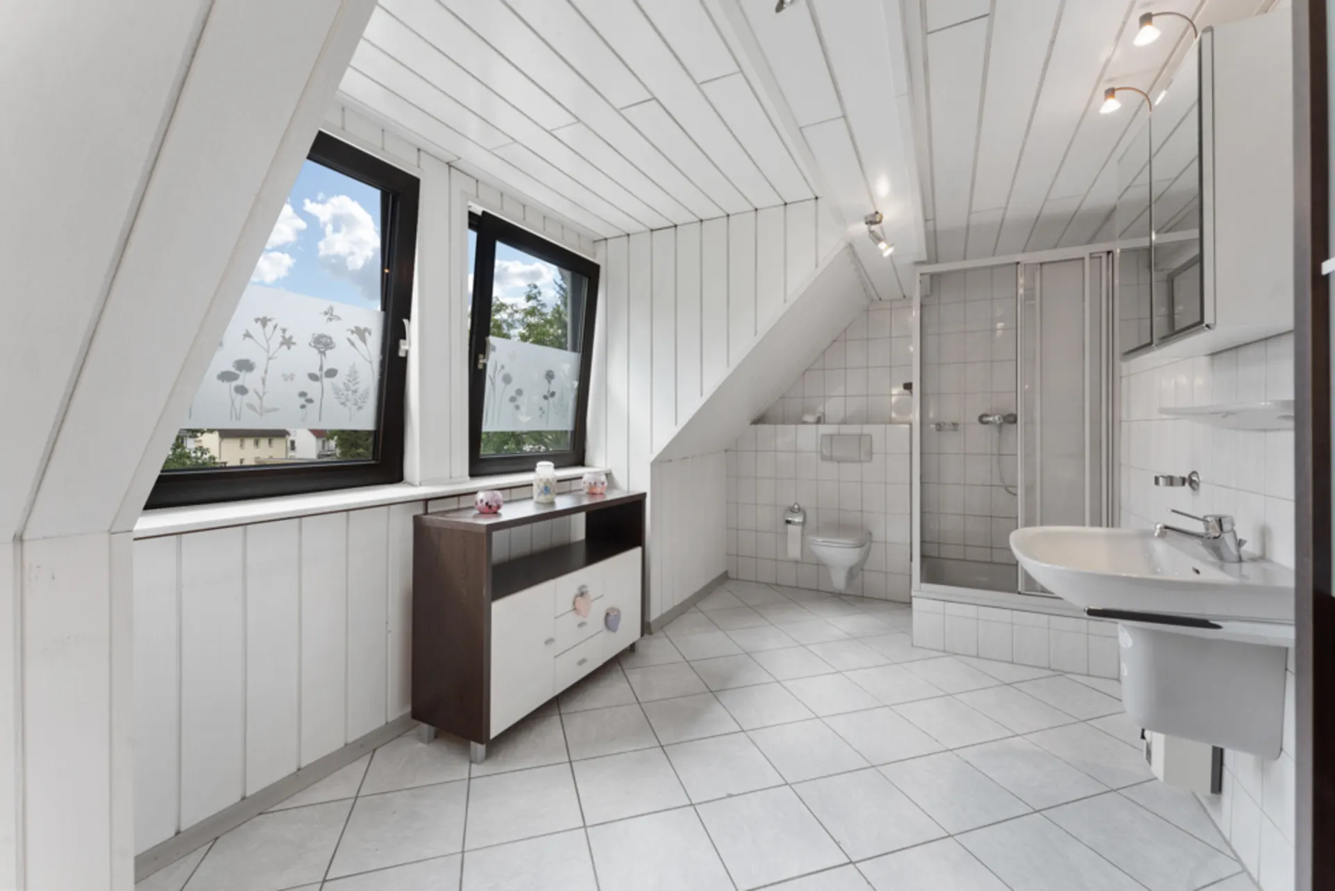 Helles Badezimmer mit Dusche, WC, Waschbecken und zwei Fenstern mit Blumenmuster-Folie.