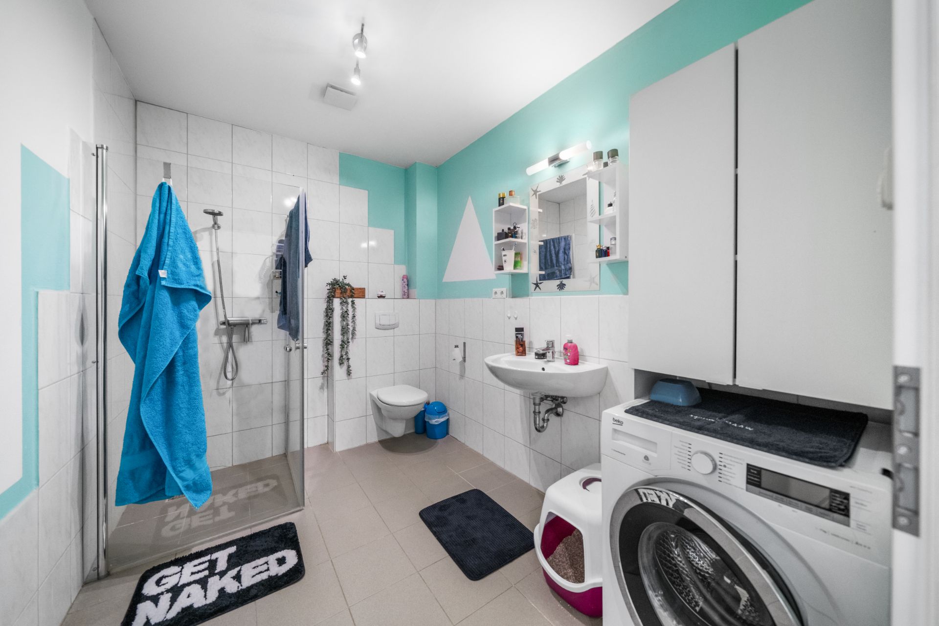 Modernes Badezimmer mit Dusche, Waschbecken, Toilette und Waschmaschine, blaue Handtücher hängen an der Wand.