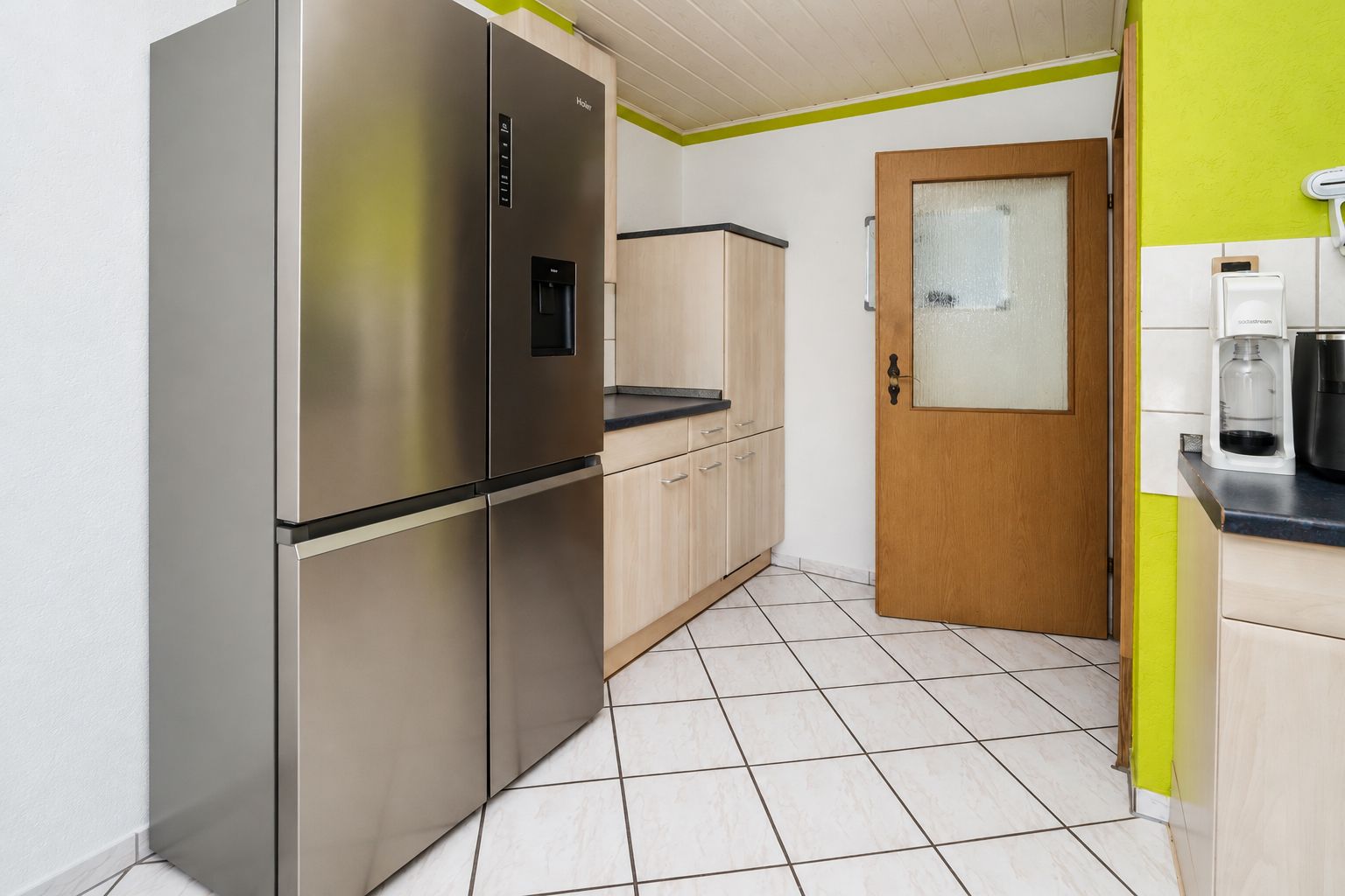 Küche mit großem Edelstahl-Kühlschrank, Holztür und hellen Fliesen.