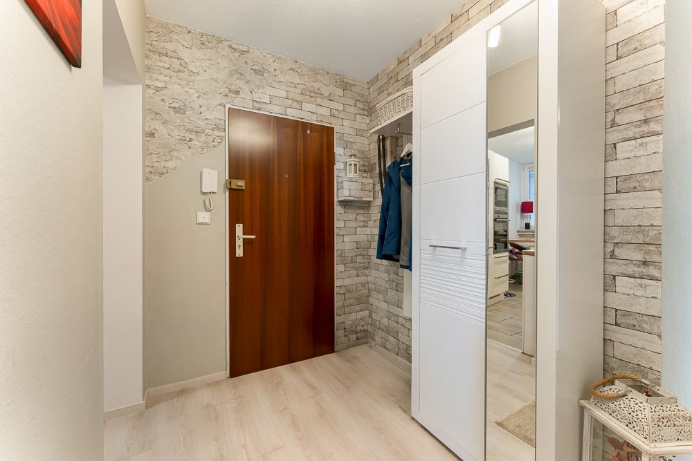 Eingangsbereich mit Holztür, Garderobe, Spiegel und hellem Boden in einer Wohnung.