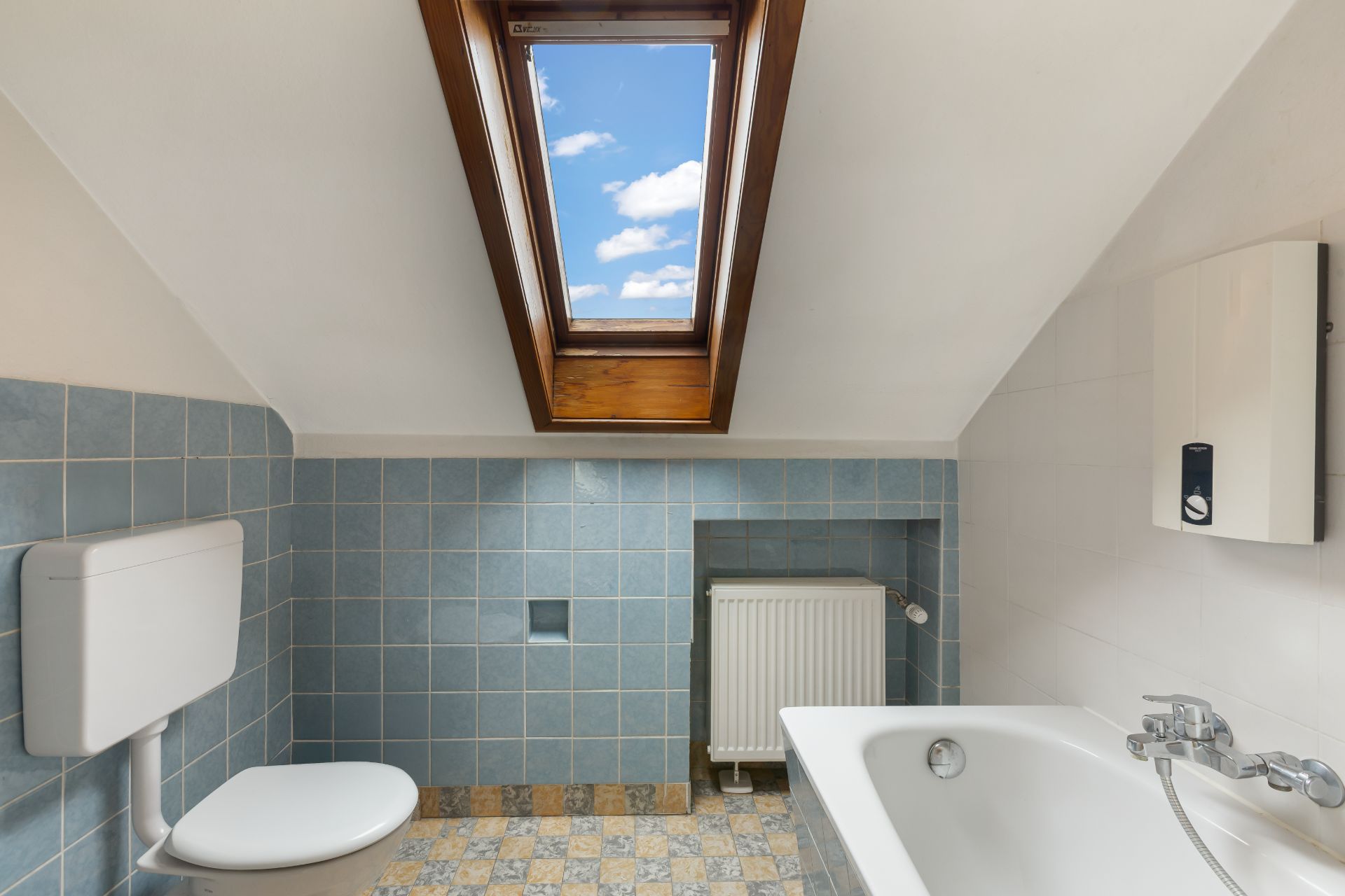 Badezimmer mit Badewanne, Toilette und Dachfenster, durch das blauer Himmel sichtbar ist.