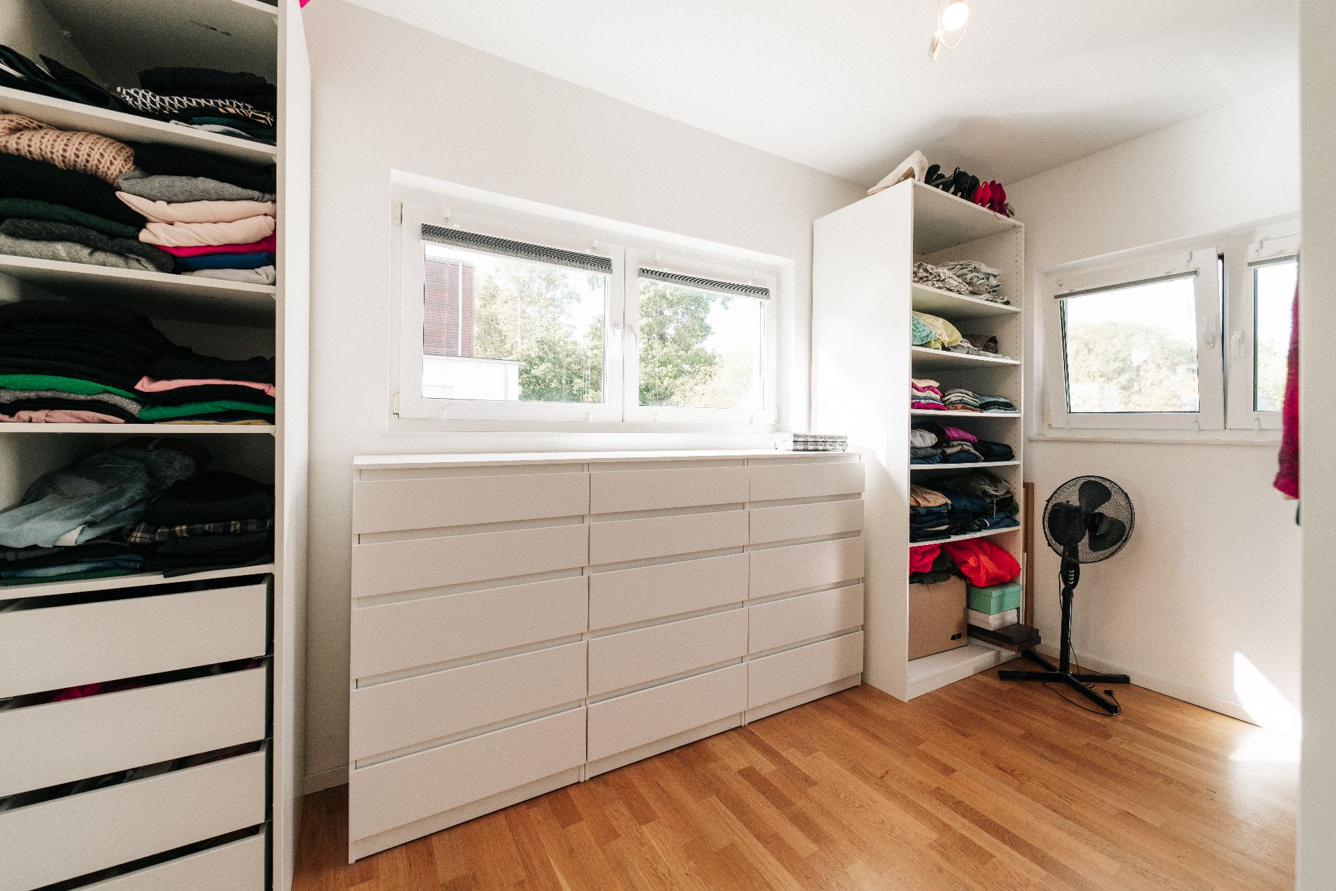 Heller Raum mit Kommode, offenen Regalen mit Kleidung und Ventilator auf Holzfußboden.