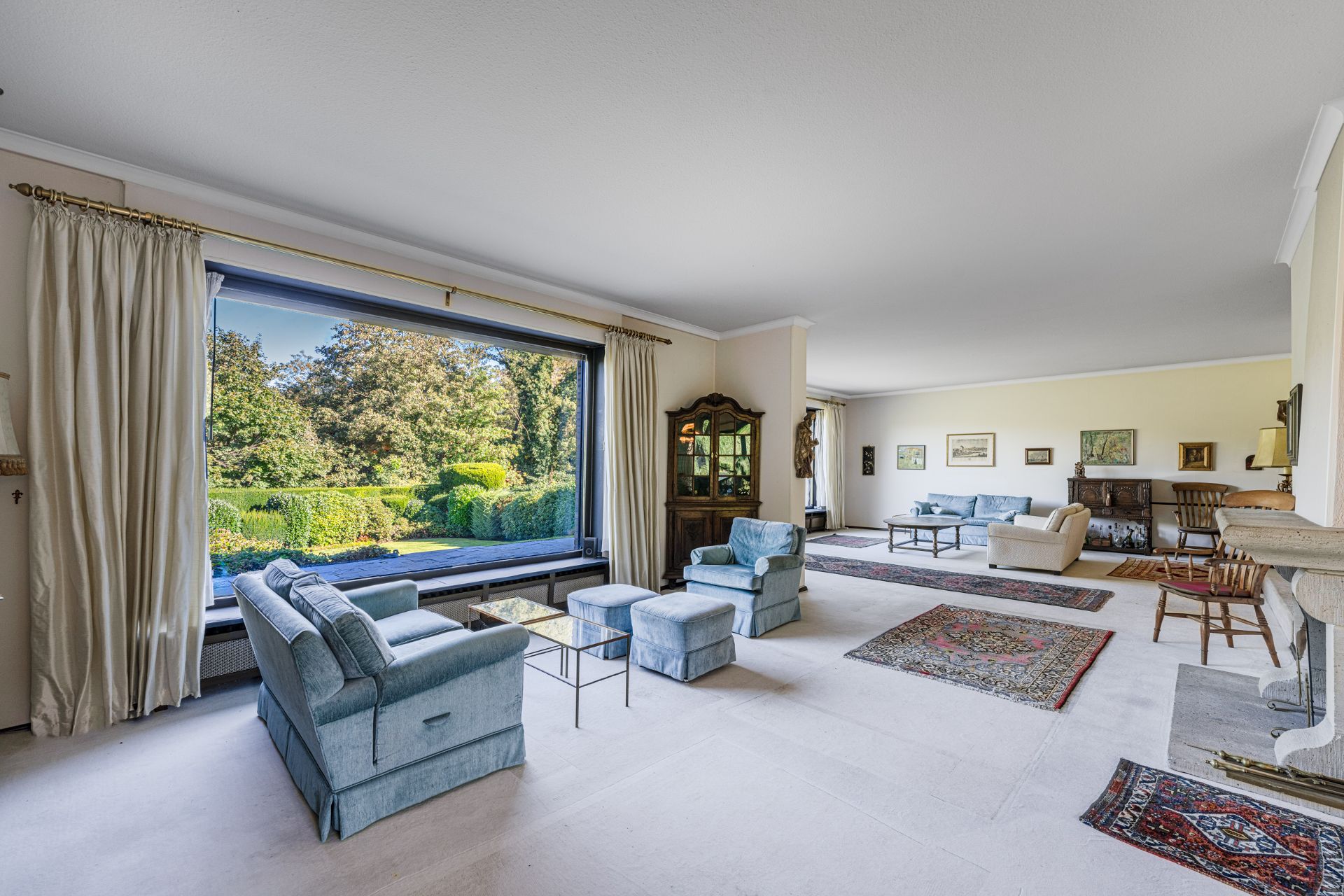 Helles Wohnzimmer mit großen Fenstern, blauen Sesseln und Blick auf einen Garten.