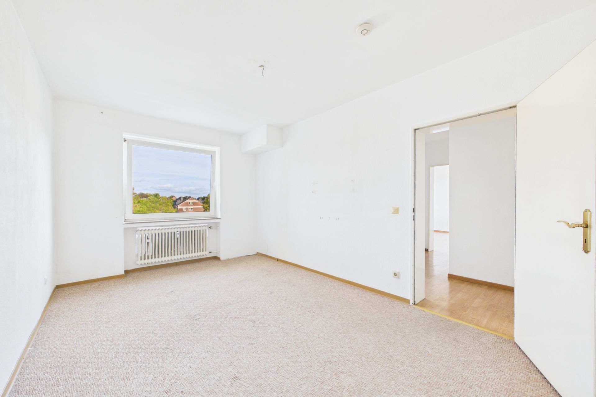Helles, leeres Zimmer mit Teppichboden, Fenster und Heizkörper, offene Tür zum Flur.
