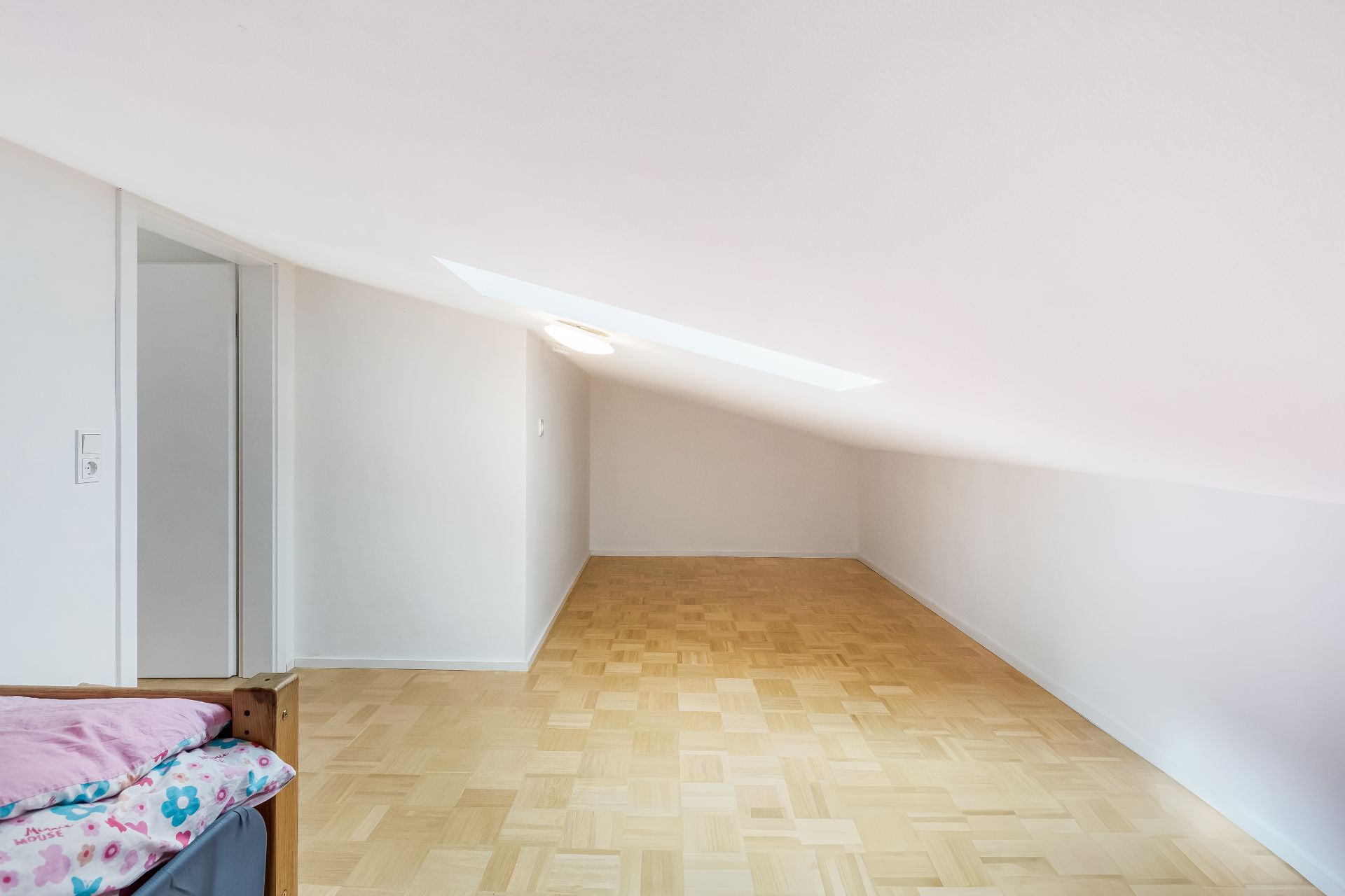 Helles Zimmer mit schräger Decke, Parkettboden und Dachfenster, im Vordergrund ein Bett mit bunter Bettwäsche.