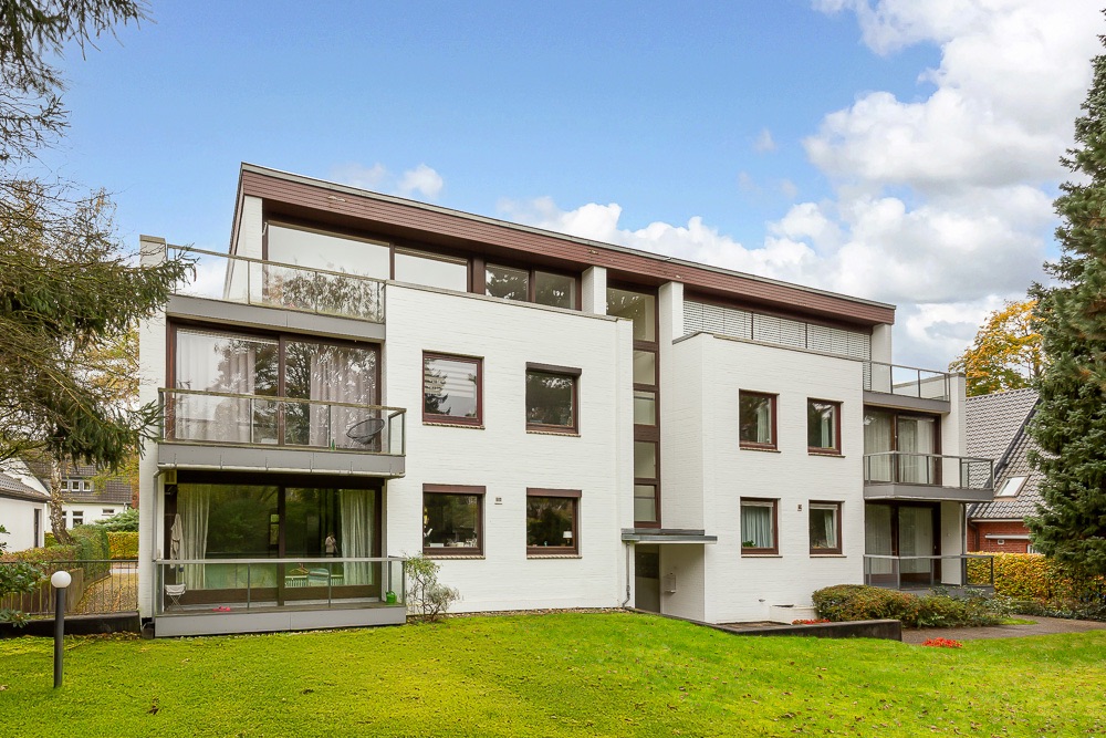 Modernes, weißes Mehrfamilienhaus mit großen Fenstern und Balkonen vor grünem Rasen.