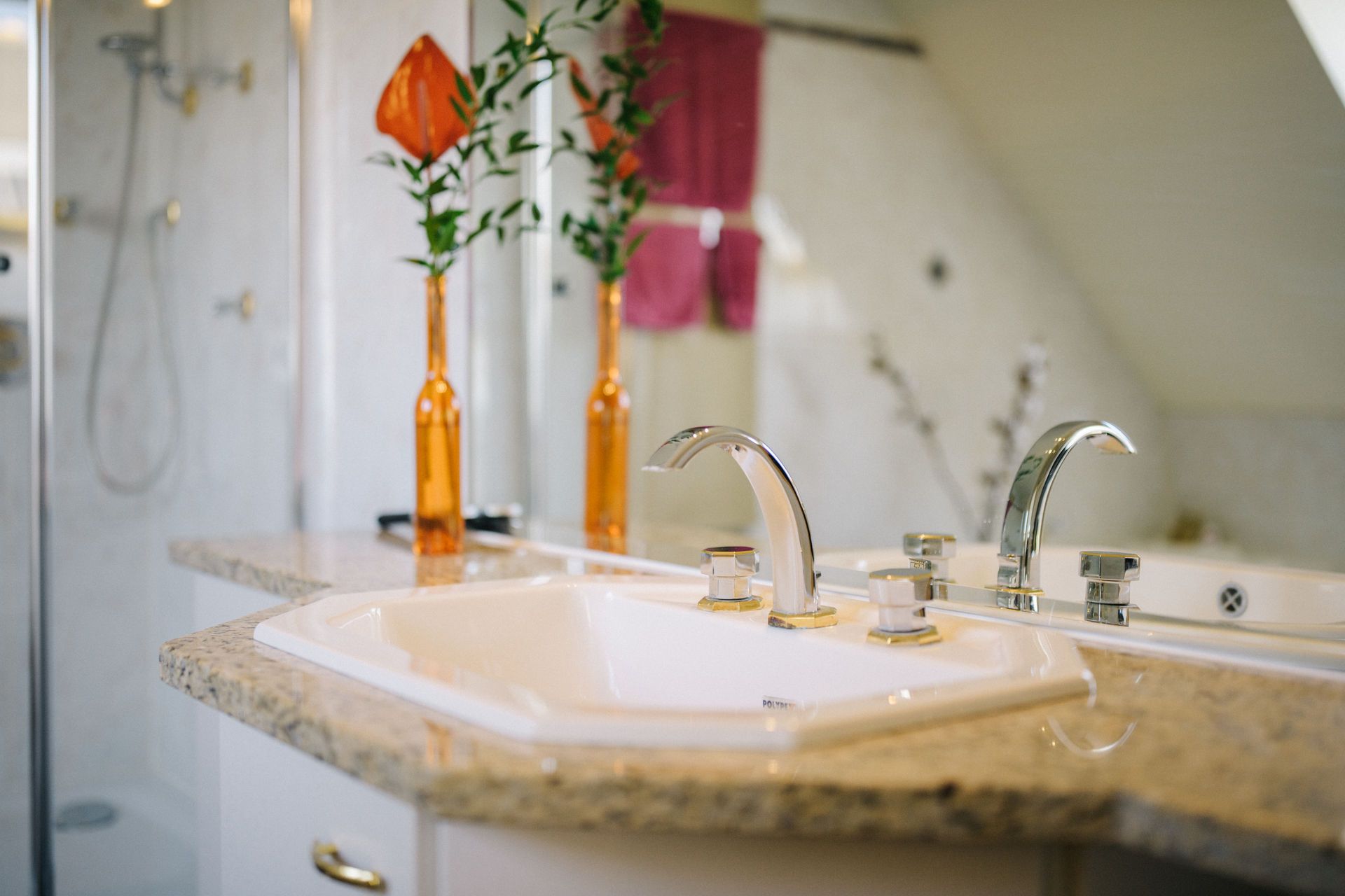 Waschbecken mit zwei Wasserhähnen und zwei orangefarbene Vasen auf einer Granitplatte im Badezimmer.