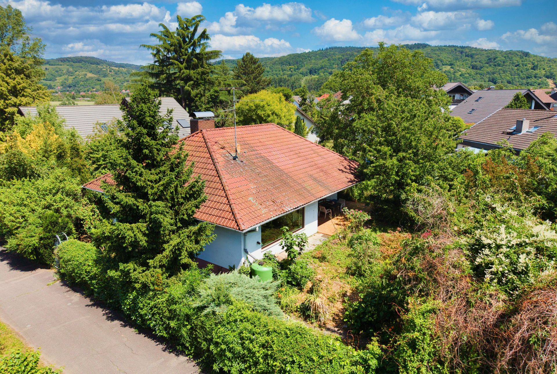 Einfamilienhaus mit rotem Dach, umgeben von Garten und Bäumen in einer grünen Wohngegend.