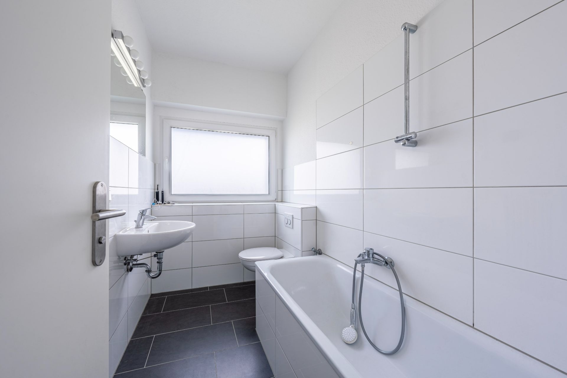 Helles Bad mit Badewanne, Waschbecken, WC und großem Fenster, weiße Fliesen und dunkler Boden.