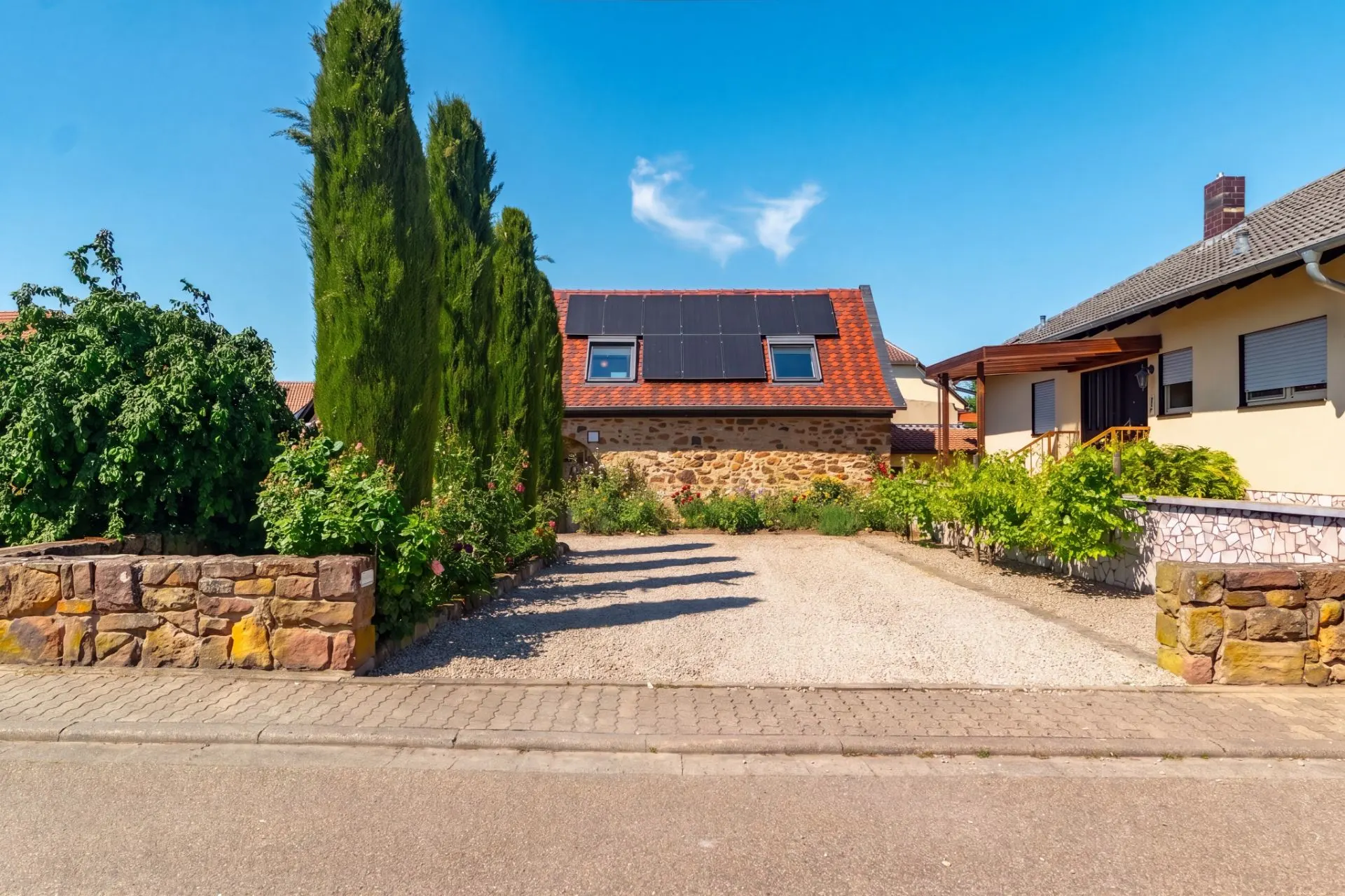Zufahrt mit Kies und Steinmauern, dahinter ein Haus mit Solarpanelen auf dem Dach.