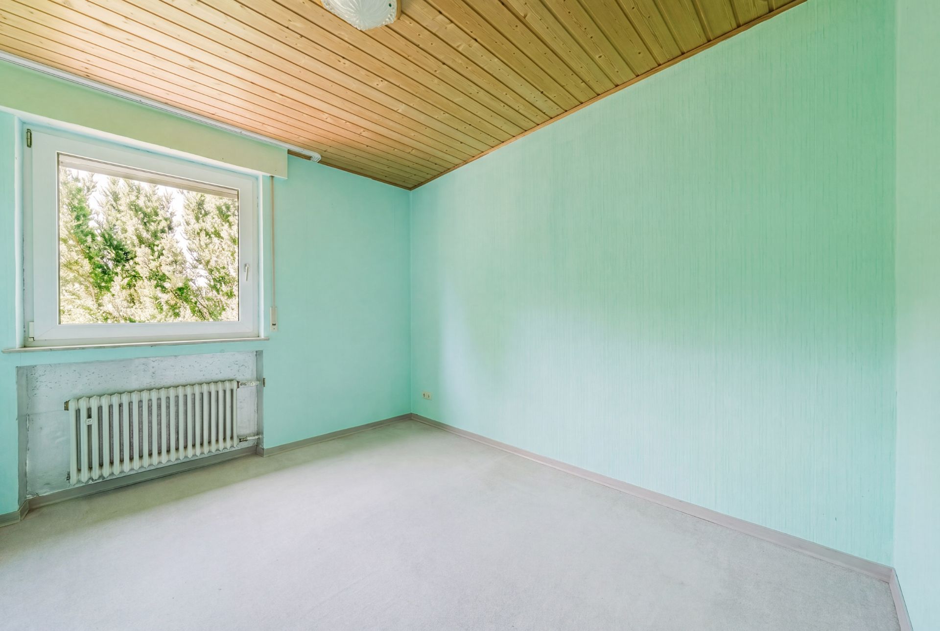 Leeres Zimmer mit mintgrünen Wänden, Holzbalkondecke und großem Fenster mit Blick ins Grüne.