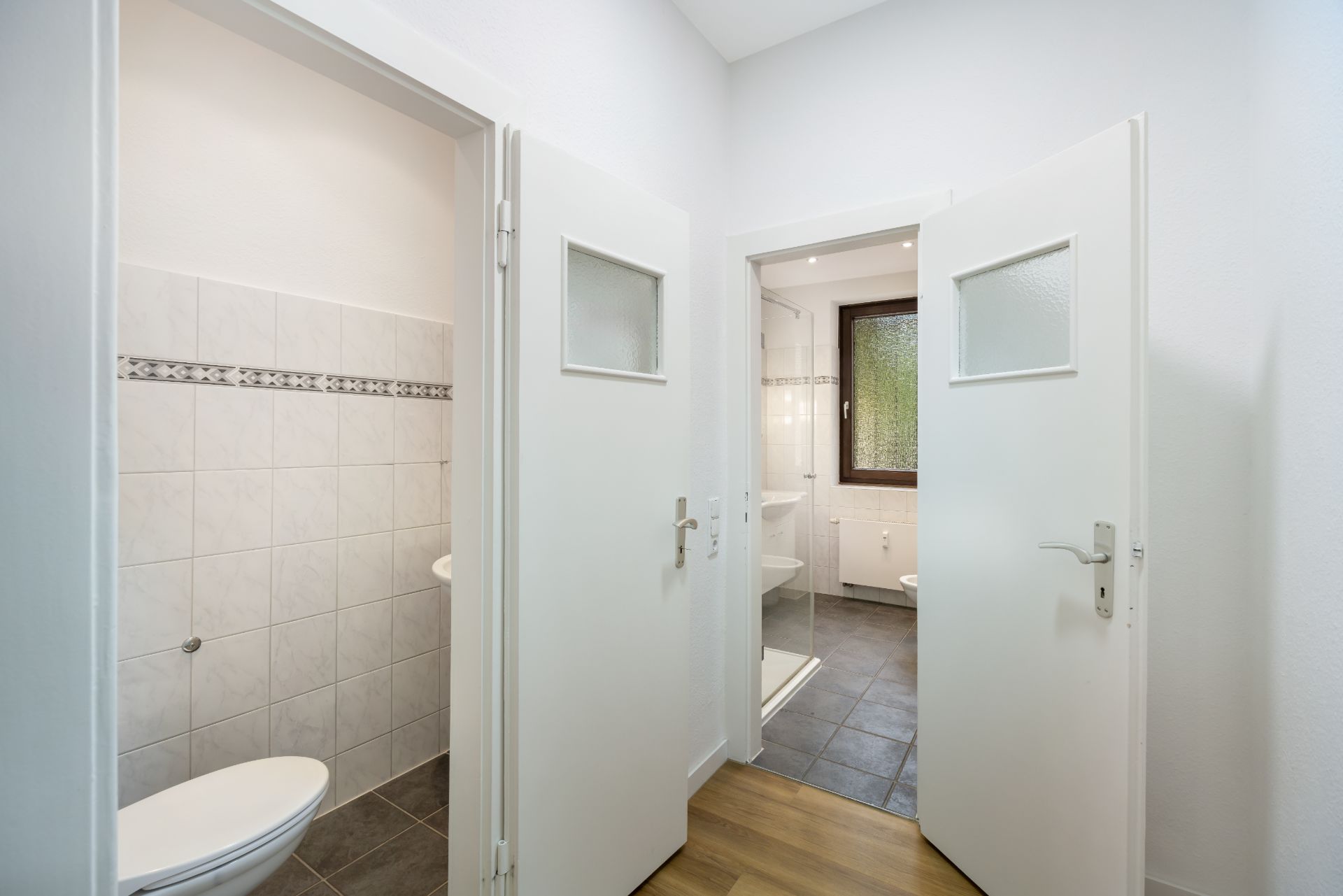 Flur mit zwei offenen Türen zu gefliesten Bädern, eines mit Fenster, Waschbecken und Toilette.